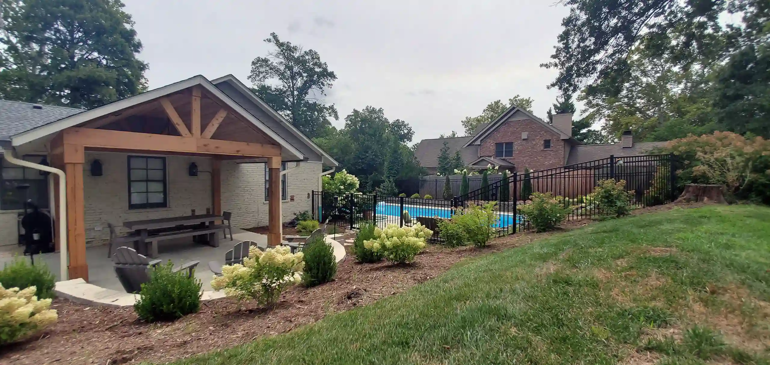 A backyard featuring a covered patio with a wooden frame, surrounded by lush landscaping and a pool area with a black metal fence.