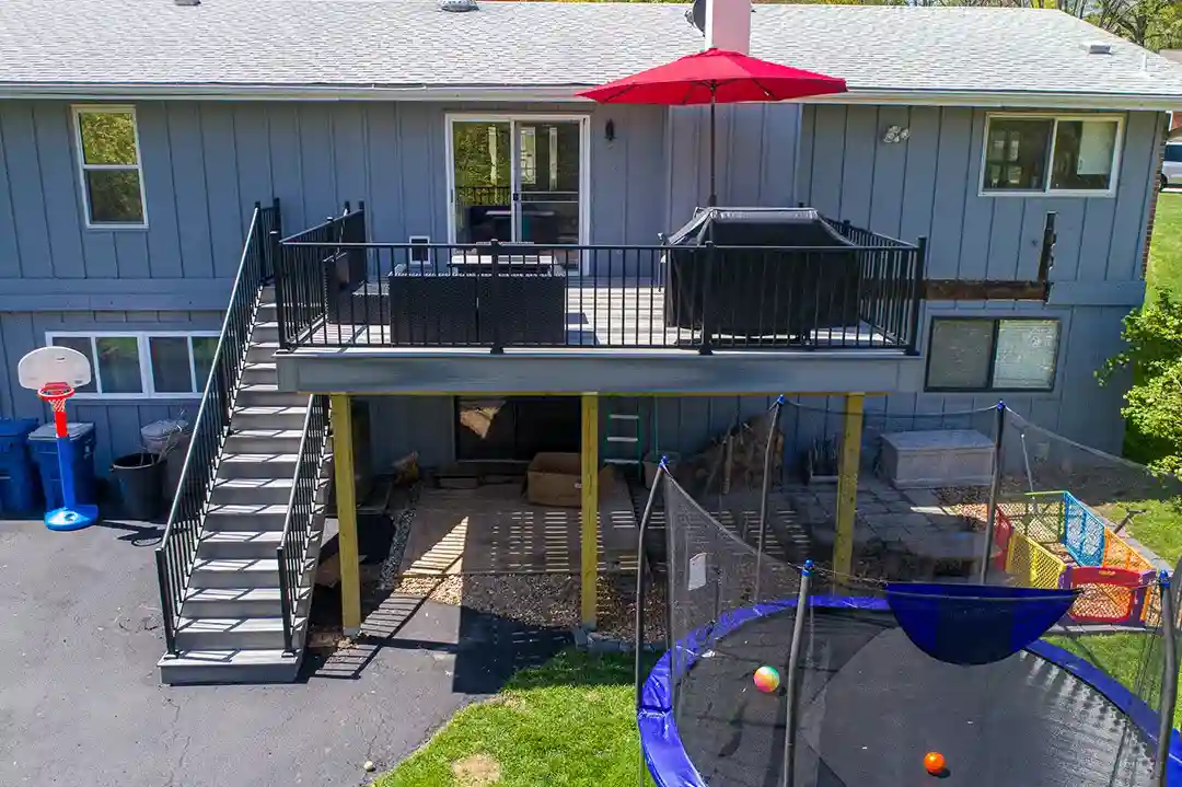 An elevated deck with black railing, a red umbrella, and a set of stairs leading down to a backyard with a trampoline and play area.

