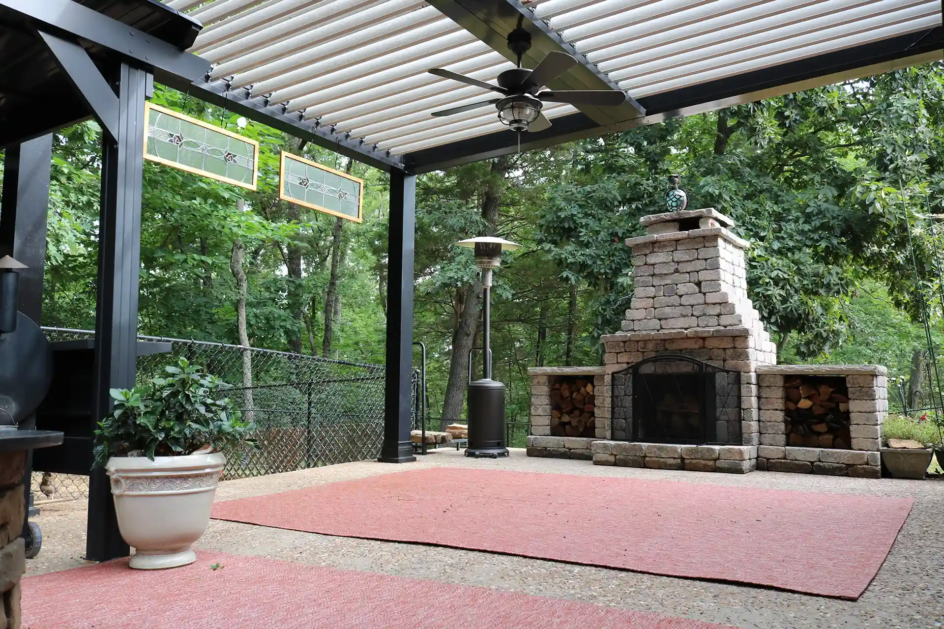 A covered outdoor living space featuring a pergola with adjustable slats, a stone fireplace, a heater, and potted plants, surrounded by trees.
