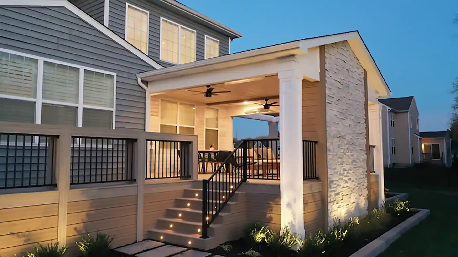 A well-lit covered porch with stone accents, a wooden deck, black railing, and a stairway.
