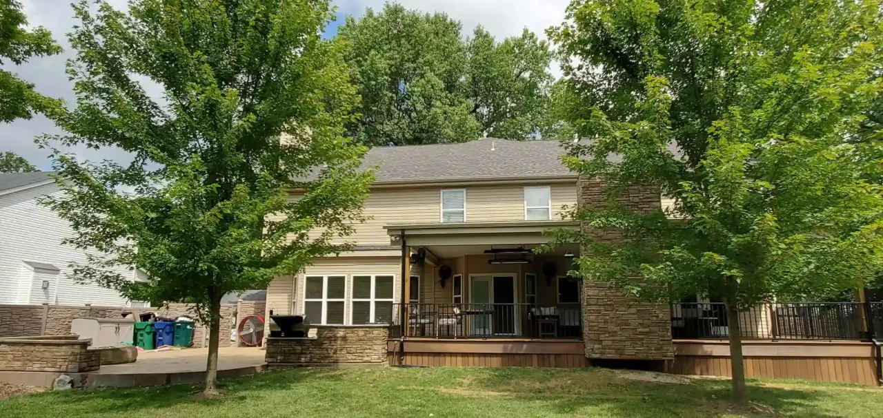 A large house with a deck, stone features, and trees in the yard.