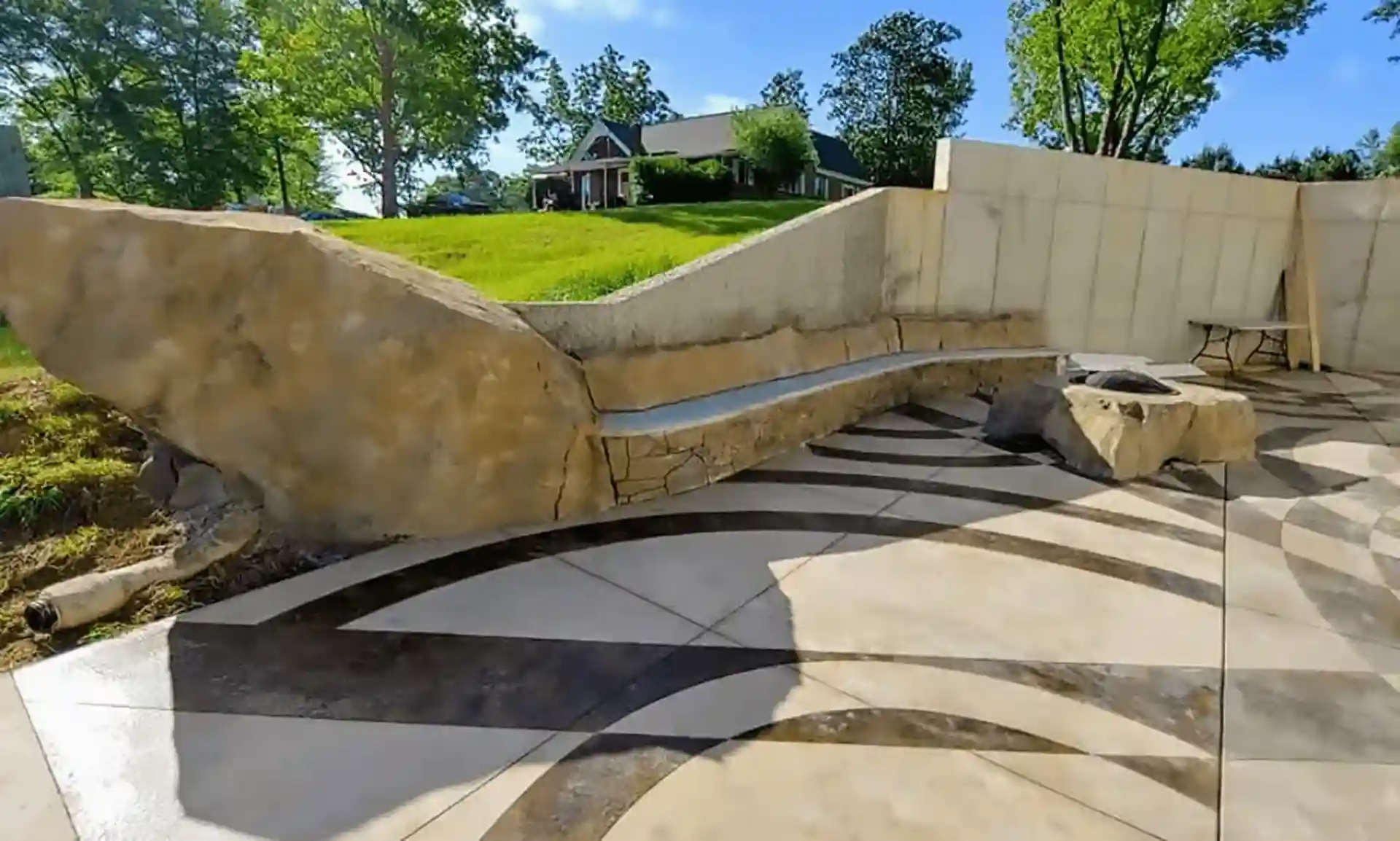 A stone bench and fire pit on a patterned patio, with a house and green lawn in the background. 
