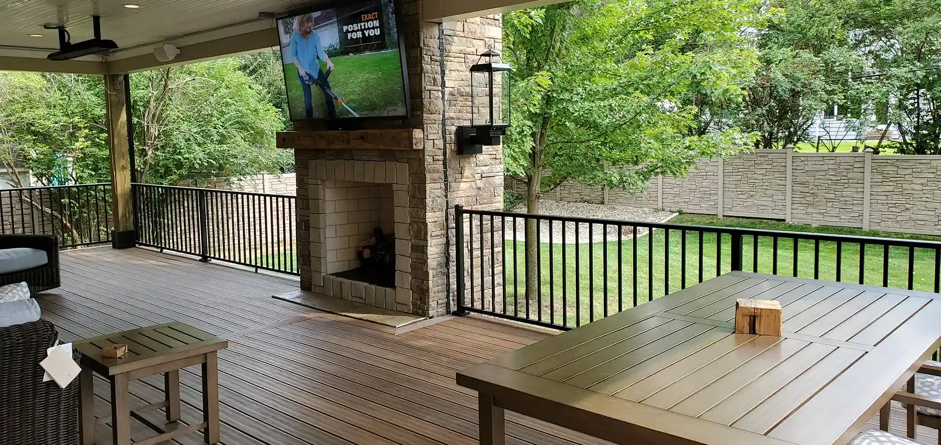 A covered outdoor deck with composite wood flooring, black railings, a stone fireplace with a TV mounted above, and a dining table. 