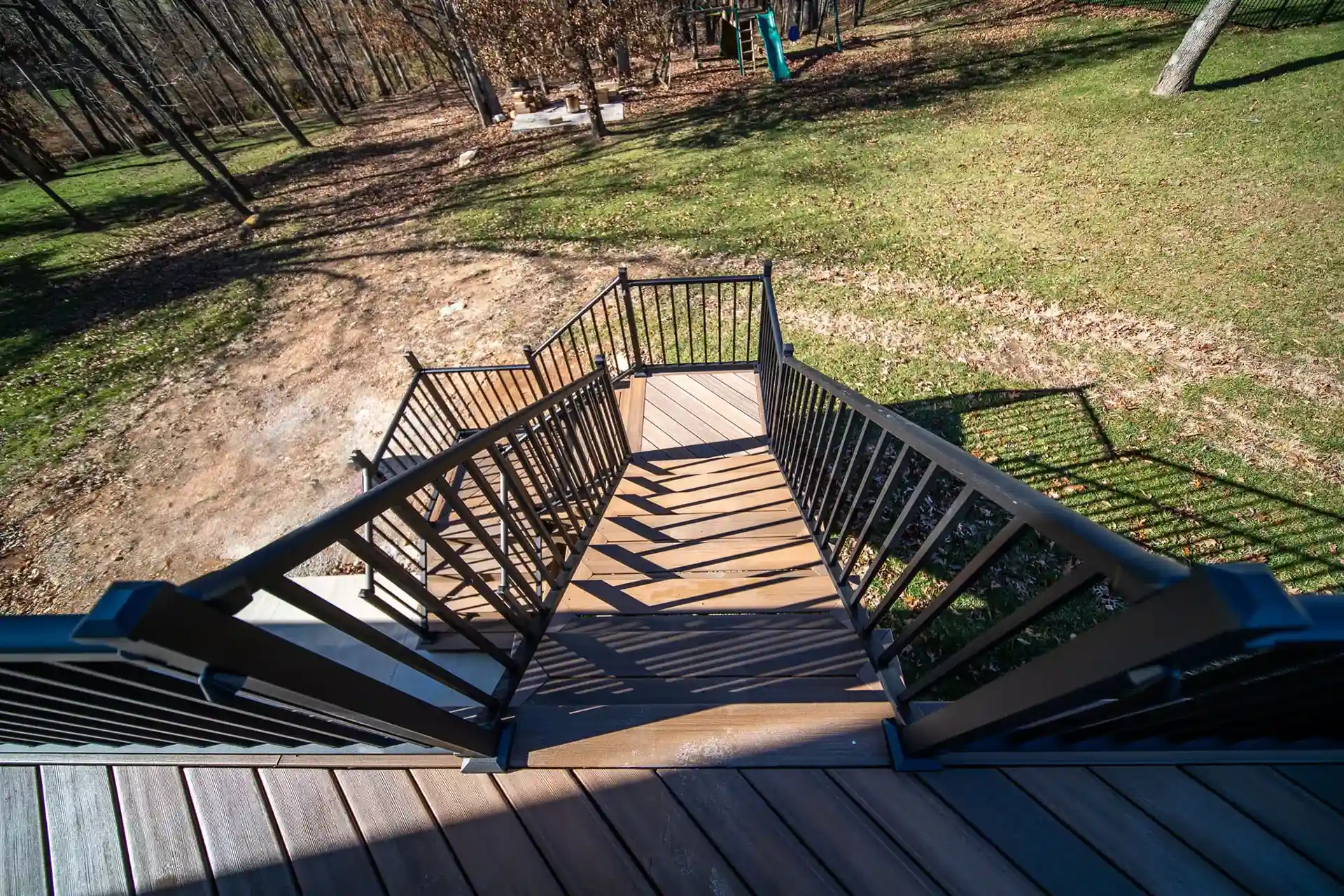A top-down view of a deck staircase with black metal railings, leading down to a grassy area with trees in the background.

