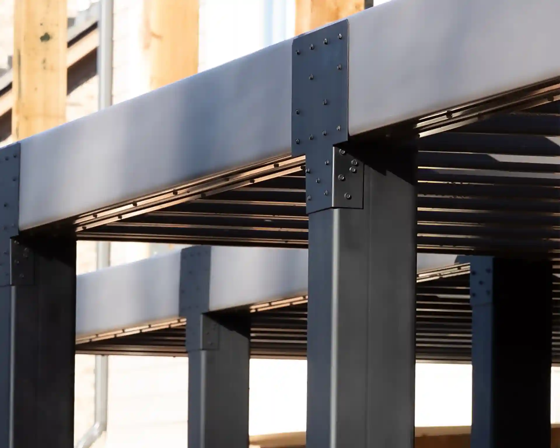 Close-up of black metal louvered pergola structure showing beams, columns, and bolted connections.
