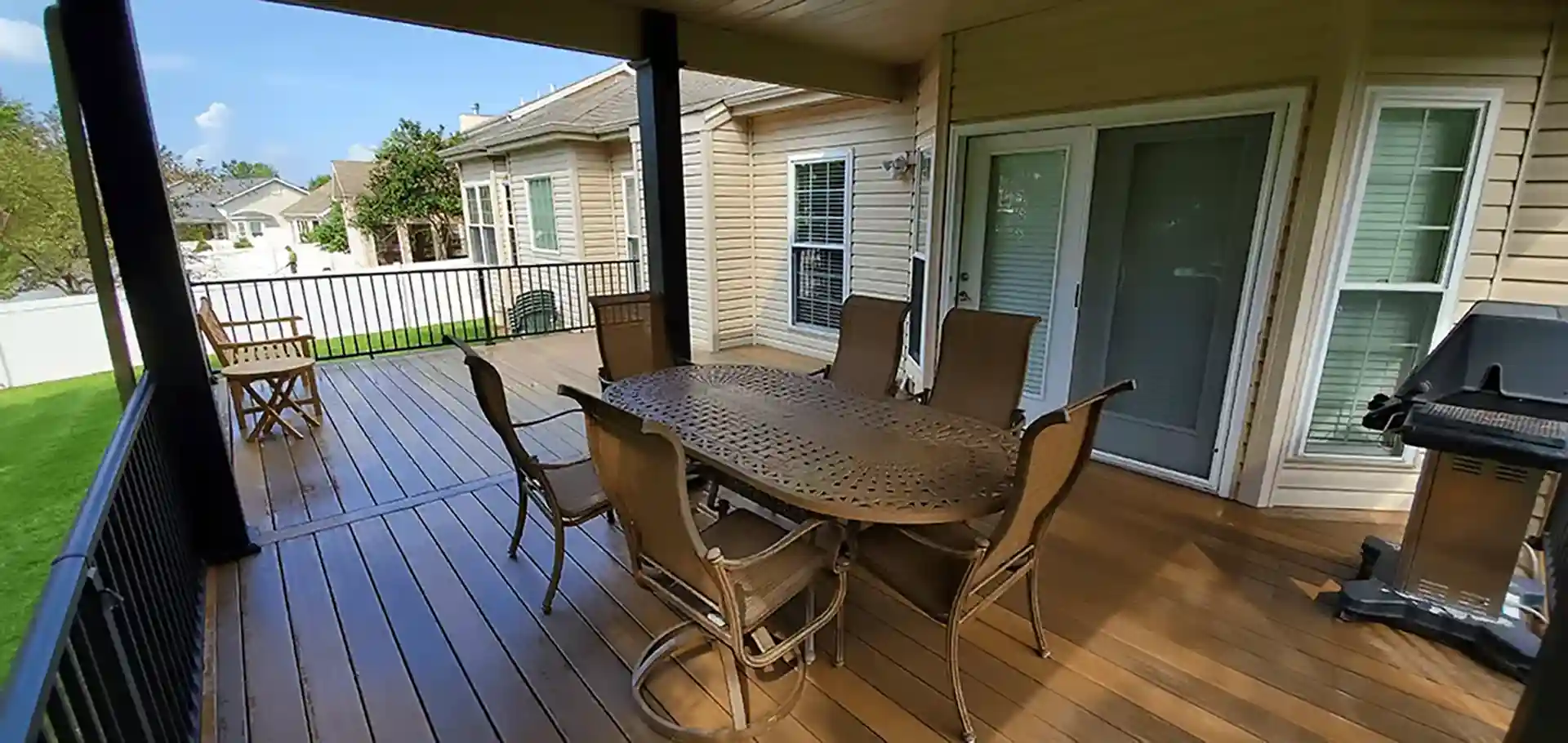 Covered outdoor deck with wood flooring, metal dining table and chairs, and a grill, overlooking a green lawn.