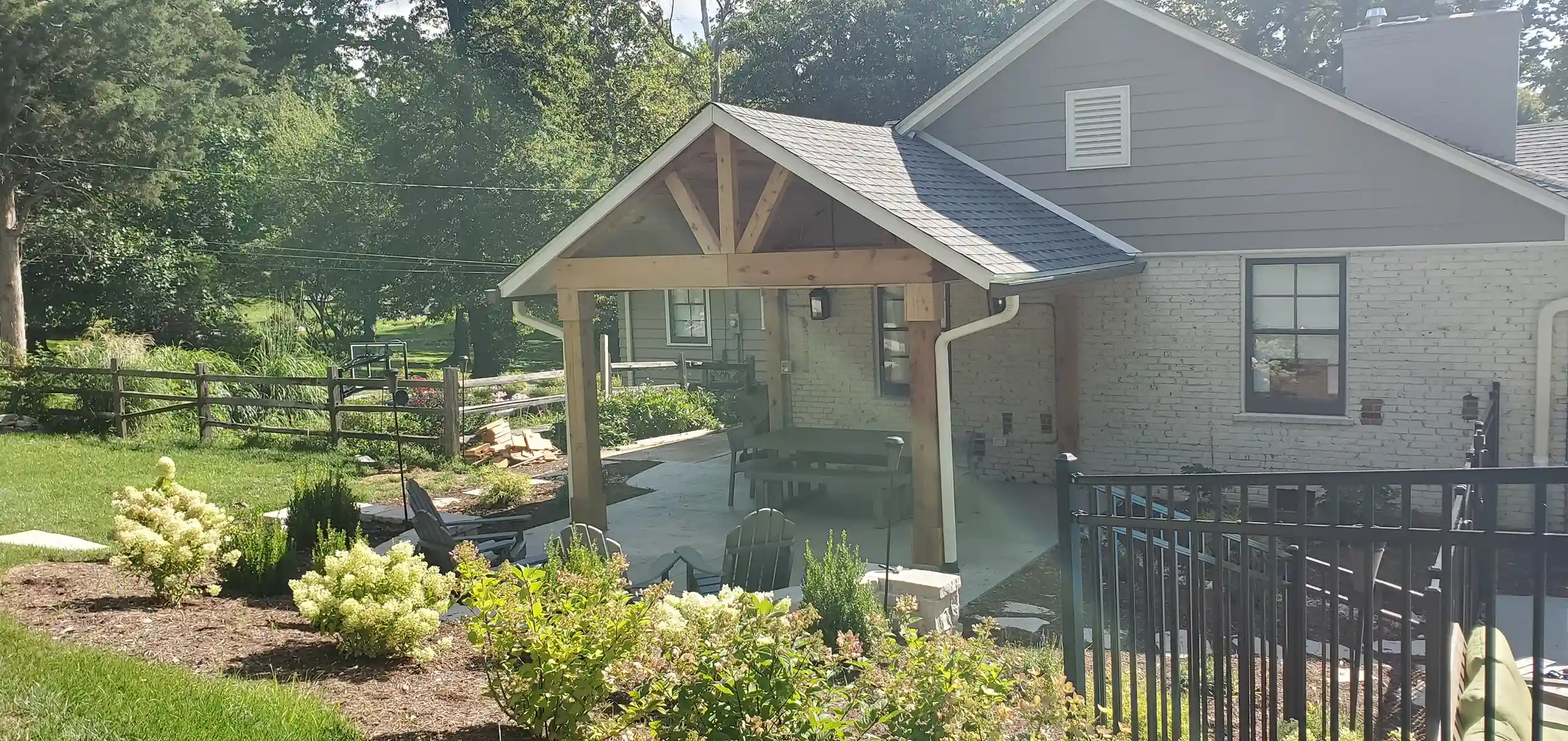 Covered outdoor patio with wooden beams and picnic table, surrounded by garden and black metal fence.