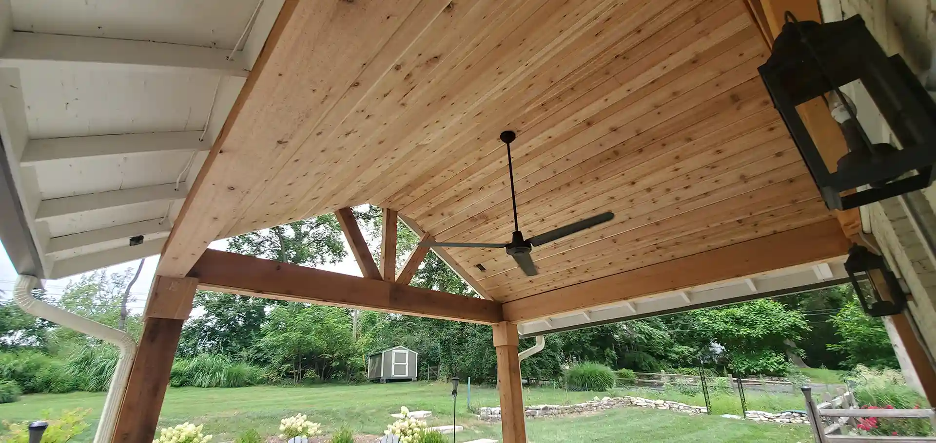 Wood-paneled covered patio ceiling with a black ceiling fan and outdoor lanterns, overlooking a green backyard with trees and garden. 