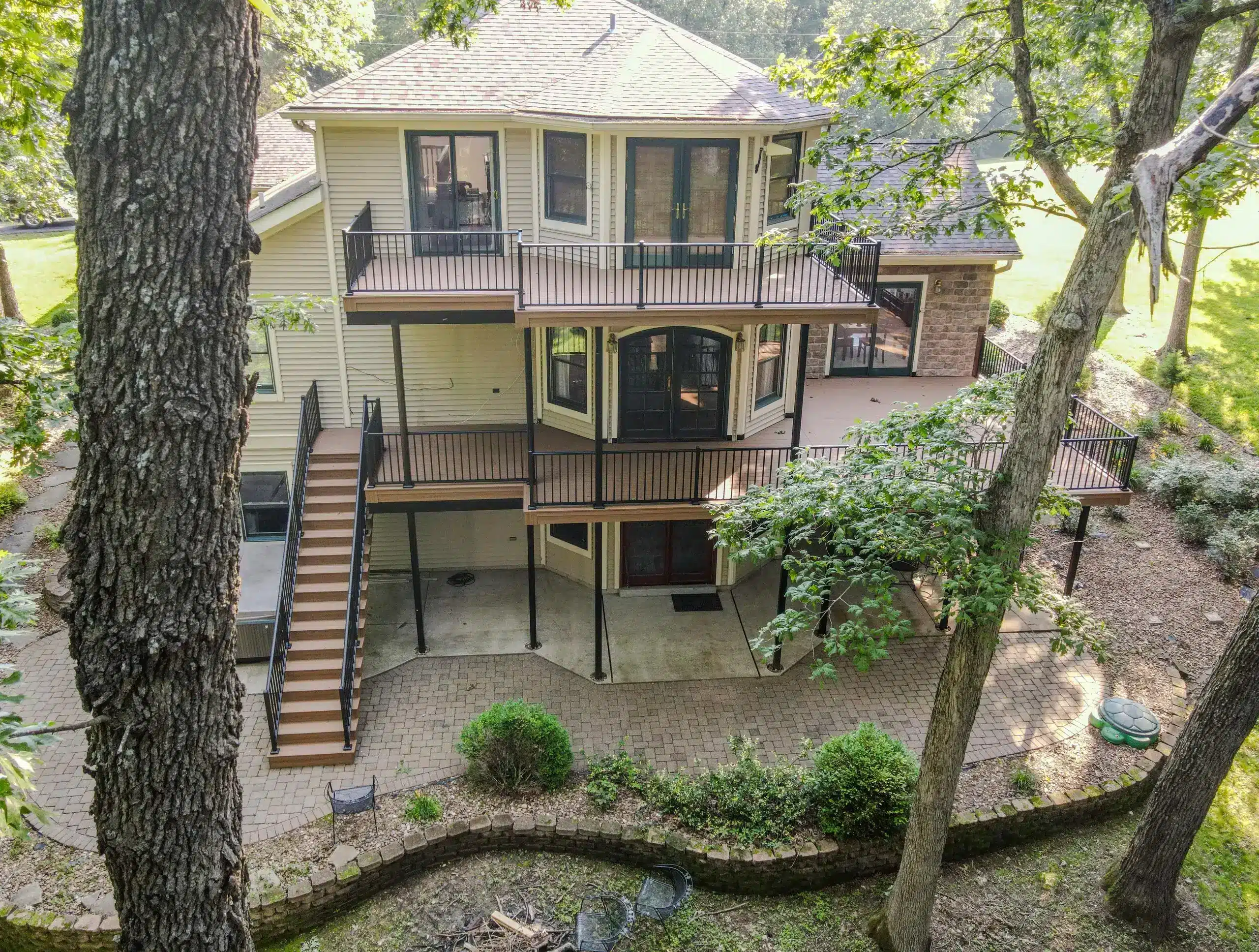 A house with two levels of decks, black railings, and a staircase leading to a ground-level patio.