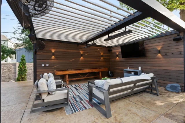 A louvered pergola covering an outdoor patio with a TV, a shuffleboard table, and seating.