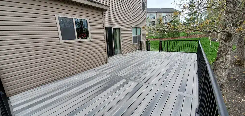 An outdoor deck made of composite material, featuring alternating light and dark gray planks. The deck is attached to a house with light brown siding, and a sliding glass door leads from the house to the deck.