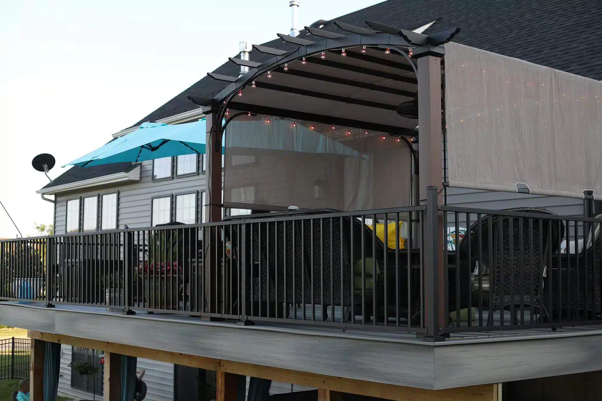 An outdoor patio with a covered pergola featuring string lights. The patio features a black metal railing and outdoor seating. In the background, there is a house with an open umbrella on an upper deck.