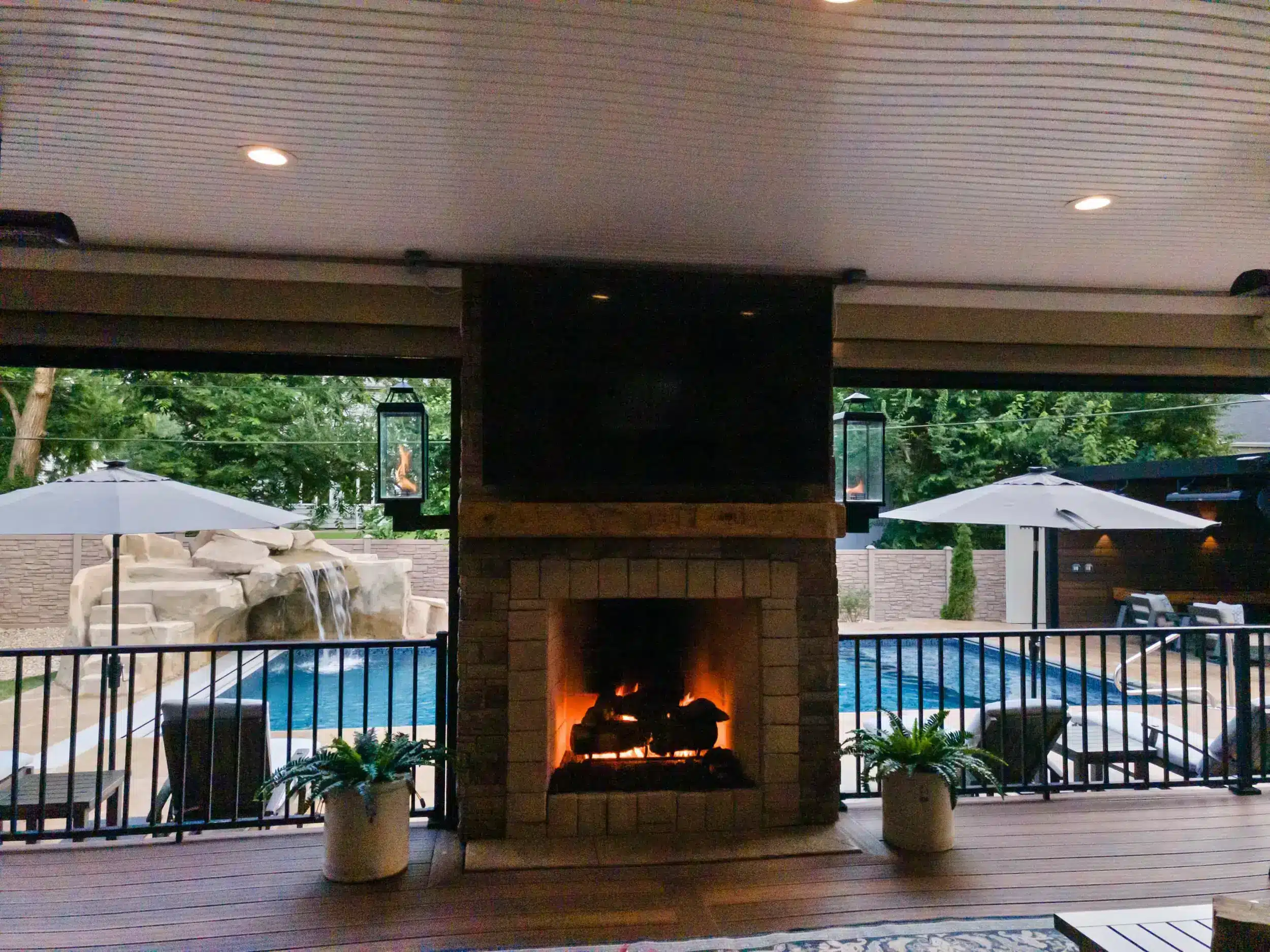 Patio Cover vs Pergola A covered outdoor living area featuring a stone fireplace with a television mounted above the mantle. A fire is burning in the fireplace. The flooring is dark wood decking. Two potted ferns sit on either side of the fireplace. Beyond the fireplace, a swimming pool is visible, surrounded by a black metal railing.