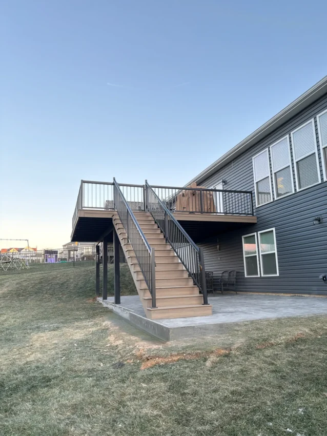 A low-angle view of a newly constructed elevated deck and staircase attached to the back of a two-story house with dark gray horizontal siding. The deck and stairs are made of light brown composite decking with a darker brown fascia and trim, and feature a black metal railing with thin vertical balusters.