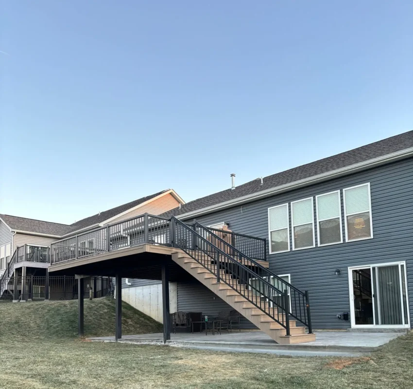 A low-angle view of a newly built elevated light brown composite deck and wide stairs attached to a dark gray-sided house. The deck has a black metal railing. The stairs lead to a concrete patio, and the deck connects to a similar structure on a neighboring house.