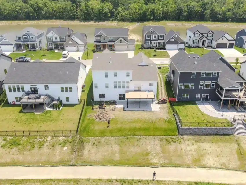 An aerial image showing the backyards of three large, modern suburban homes. The central white house is undergoing construction with a newly poured covered patio, an excavator, and two people in the yard. The backyards are separated by fences, and a wide cleared area slopes down to a path and a body of water, with more houses visible across the water.