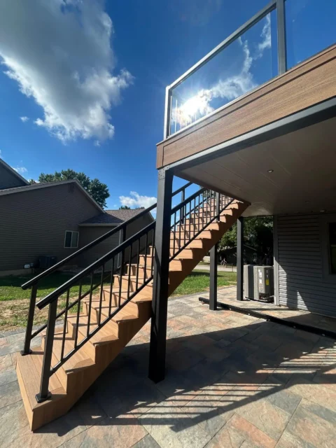 A compsoite staircase with black metal railings leads up to a second-level deck with a clear glass railing, next to a house with gray siding under a sunny sky.