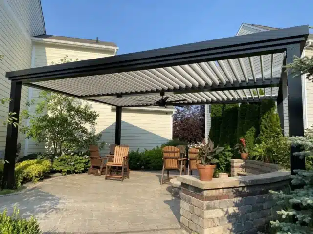 A black-framed louvered aluminum pergola covers a stone patio with wooden outdoor chairs and potted plants, situated between a light-colored house and landscaping.