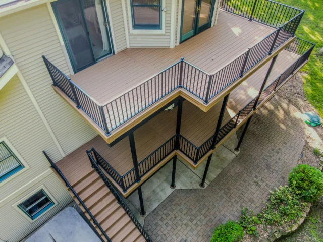 A two-story residential deck with tan composite decking and black metal railings is shown. The deck features a staircase leading down to a paved patio area.