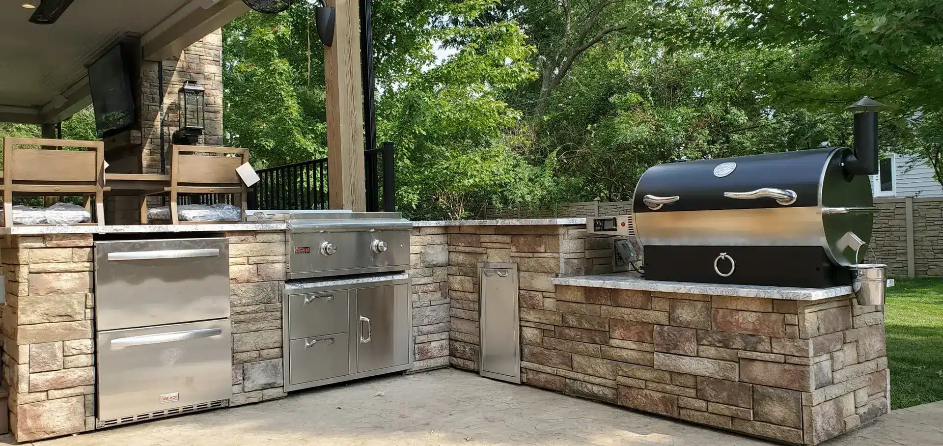 An outdoor kitchen is depicted in the image. The kitchen features stone masonry surrounding built-in stainless steel appliances, which include a large grill or smoker with a chimney, a drop-in gas grill, a refrigerator, and what appears to be a narrow storage unit or trash pull-out.