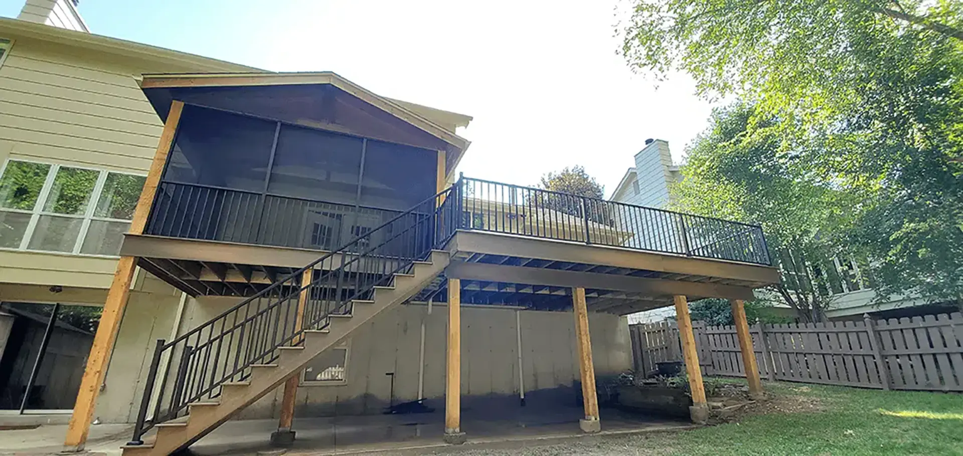 A two-story deck structure attached to the rear of a light-colored house. The deck features a screened-in porch area on the upper level and an open deck section, both enclosed by black metal railings. A set of stairs with black metal railings descends from the open deck to the ground. The deck is supported by several light-brown or natural-wood posts.